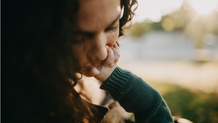 Anschnitt eines Frauenkopfes mit dunklen Locken nach rechts blickend. Sie ist leicht vonr über gebeugt. Ein Babyarm kommt von unten rechts, die Hand fasst der Frau in an den Mund und greift ihre Lippen